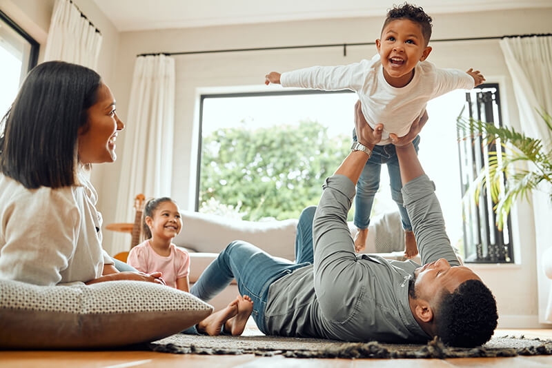 A joyful family plays together in a bright living room, with a child being lifted by a parent while two others watch and smile.